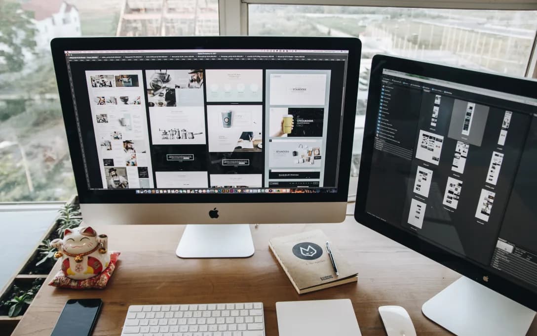 Two desktop monitors showing web page layouts and design boards on a designer's desk