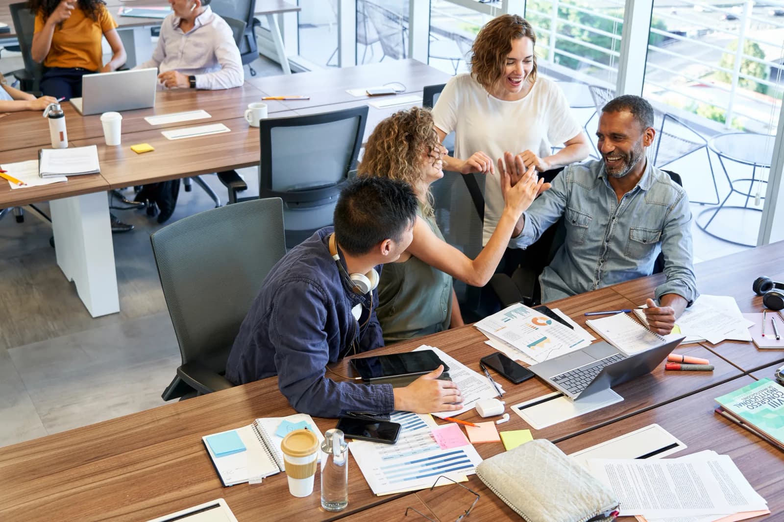 Four colleagues high-fiving across a meeting table covered with printed charts, laptops and sticky notes