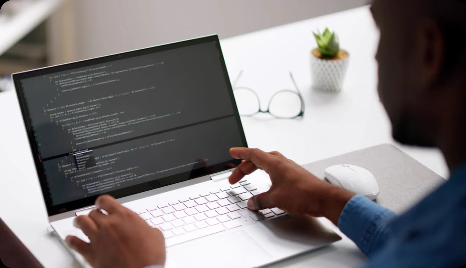 A developer typing on a laptop showing lines of code, with glasses and a plant on the desk